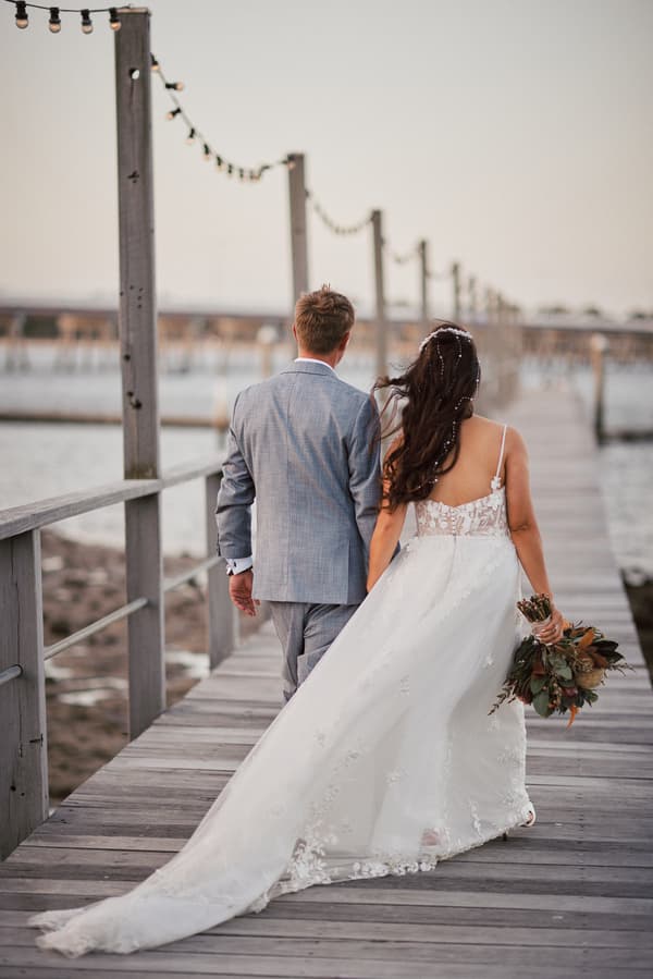 The bride Jheng in a white wedding gown holding a bouquet and the groom Dane in a light gray suit walk hand in hand along a wooden pier at Sandstone Point Hotel.