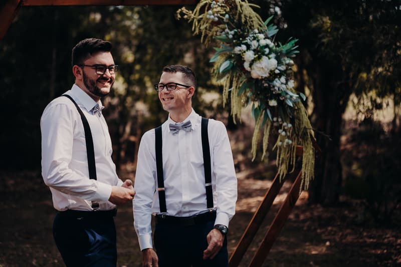 Two groomsmen in white shirts with suspenders and bow ties stand near a floral wedding arch at Ocean View Estates.