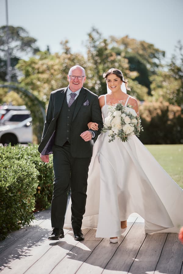 The bride Ashleigh walks arm-in-arm with an older man, likely her father, along a wooden pathway outdoors at Sandstone Point Hotel — The Pavilion, holding a bouquet of white flowers.