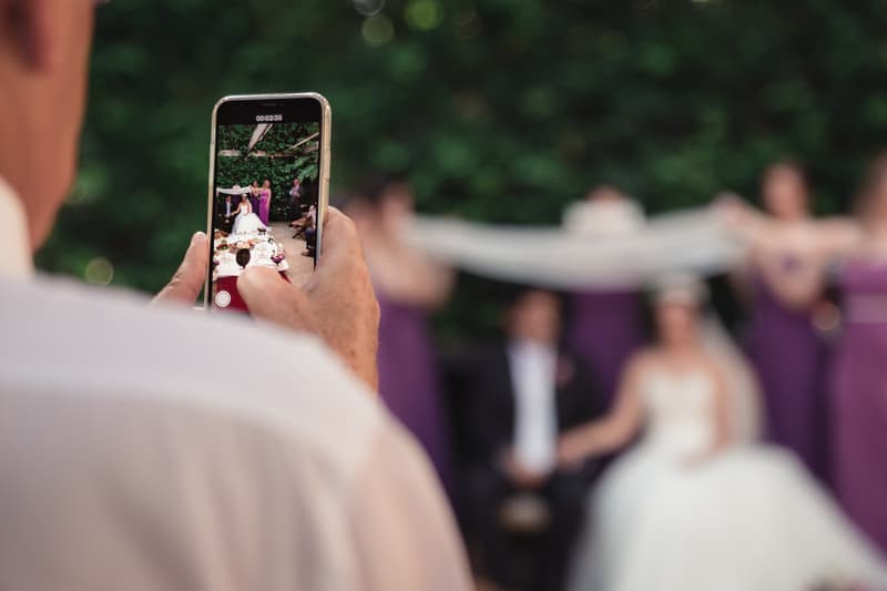 A guest captures a video of the bride Maryam, groom Pasha, and bridesmaids at Hillstone St Lucia — The Quartyard during the ceremony or formal group photo.