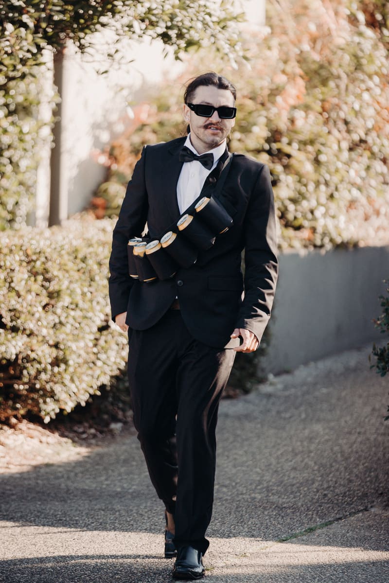 A groomsman in a black tuxedo and sunglasses walks outdoors, wearing a bandolier holding several cans. Sunset wedding portraits Brisbane by She Said Yes.