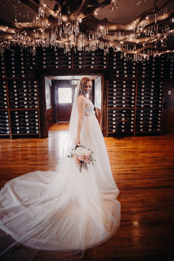 The bride Chelsea poses alone holding a bouquet in a room at Preston Peak Winery Functions Centre with wooden floors, wine racks, and a chandelier with hanging crystals.