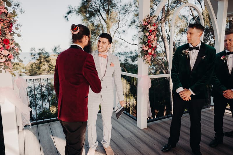 David, the groom, in a light grey suit with a peach bow tie, shakes hands with a man in a burgundy jacket on the ceremony stage at Sandstone Point Hotel — Pavilion. Two groomsmen in dark green tuxedos with black bow ties stand to the right, near floral decorations on white pillars.