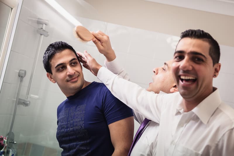 The groom is having his hair brushed by a man in a white shirt and purple tie in a bathroom with a glass shower door visible.