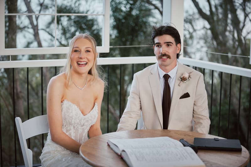 The bride Morgan and groom Joel sit at a round wooden table with an open book and a black folder during their ceremony at Sandstone Point Hotel — The Pavilion.