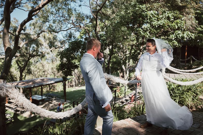 Bride Jheng in a white wedding dress and veil stands on a stone path outdoors at Sandstone Point Hotel, adjusting her veil while groom Dane in a light grey suit faces her, surrounded by trees and greenery.