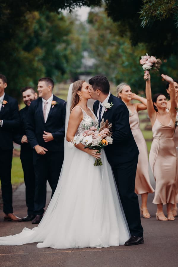 Chelsea the bride and Brent the groom kiss outdoors at Preston Peak Winery Functions Centre with bridesmaids in champagne dresses and groomsmen in dark suits standing behind them.