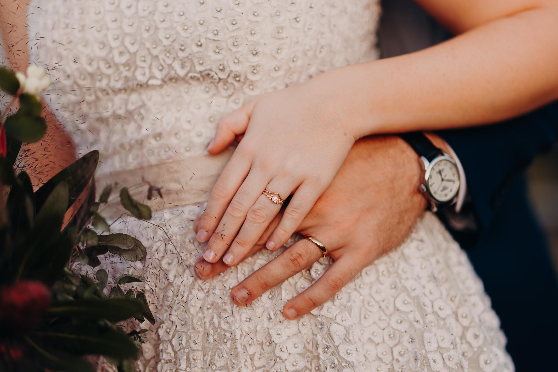 Close-up of the bride and groom's hands resting on the bride's beaded wedding dress at Yabbaloumba Retreat, showing their wedding rings and part of the bride's bouquet.