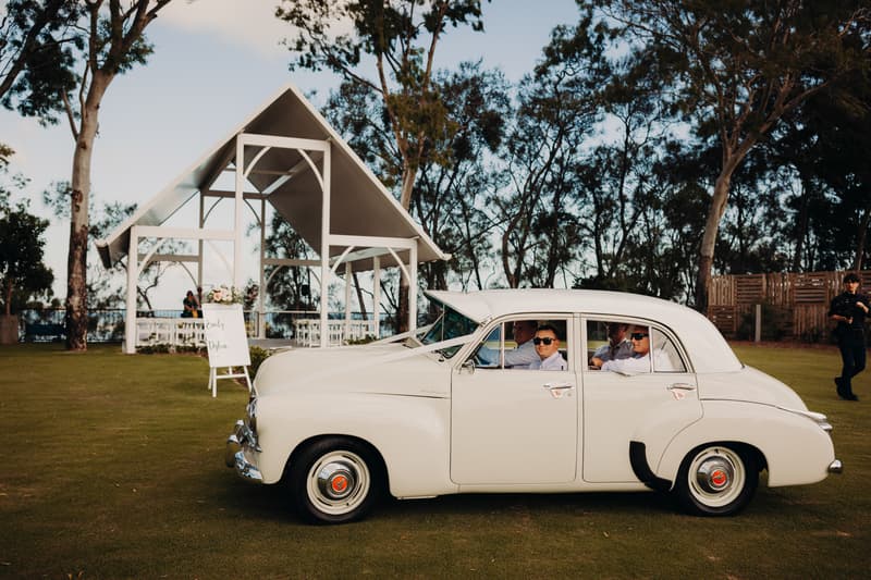 A vintage white car with four men wearing white shirts and sunglasses is parked on the grass near the ceremony pavilion at Sandstone Point Hotel. A wedding sign with the names Emily and Dylan is visible near the pavilion.