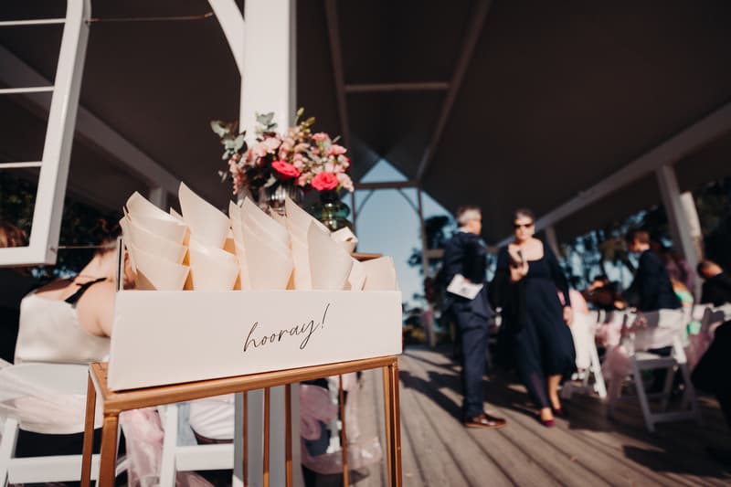 Ceremony setup at Sandstone Point Hotel — Pavilion with a box labeled 'hooray!' holding paper cones, guests seated on white chairs with pink sashes, and two guests conversing in the background.