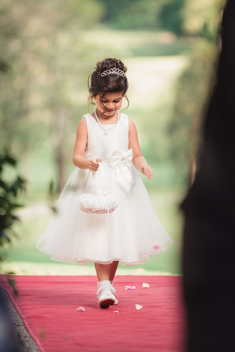 A flower girl in a white dress and tiara walks down a red carpet scattering petals. Sunset wedding portraits Brisbane by She Said Yes.