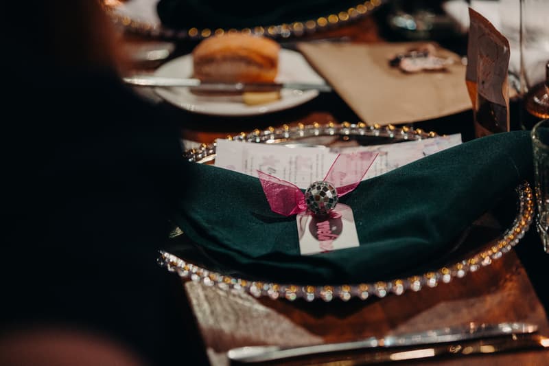 Close-up of a wedding reception place setting at Sandstone Point Hotel — Cellar, featuring a dark green napkin with a pink ribbon and a small disco ball decoration on a beaded charger plate, with a bread roll and cutlery visible in the background.