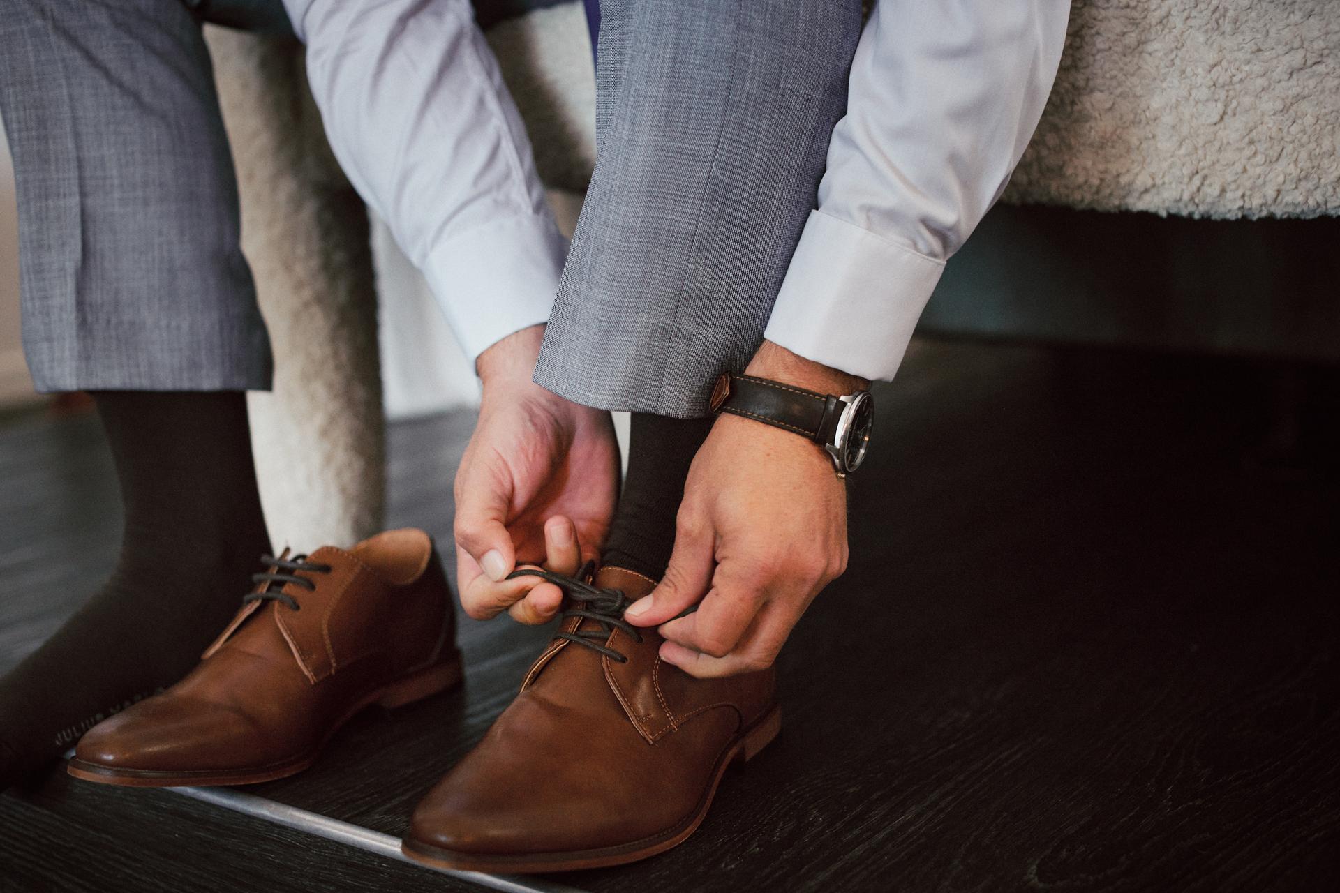 The groom at Sandstone Point Hotel is tying the laces of his brown dress shoes while seated on a bed.