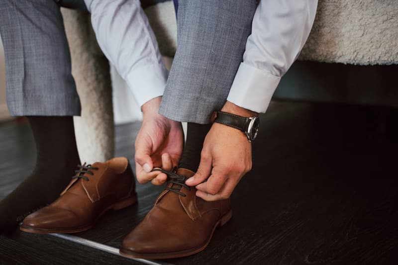 The groom at Sandstone Point Hotel is tying the laces of his brown dress shoes while seated on a bed.