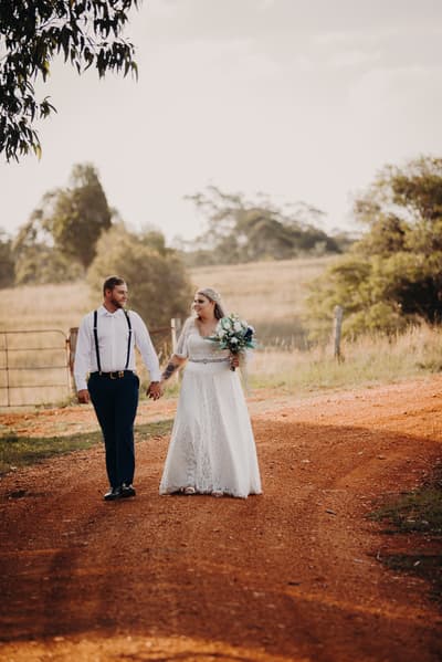 Annie and Trent walk hand in hand on a dirt path outdoors, with Annie holding a bouquet of flowers. Photo and video services by She Said Yes.