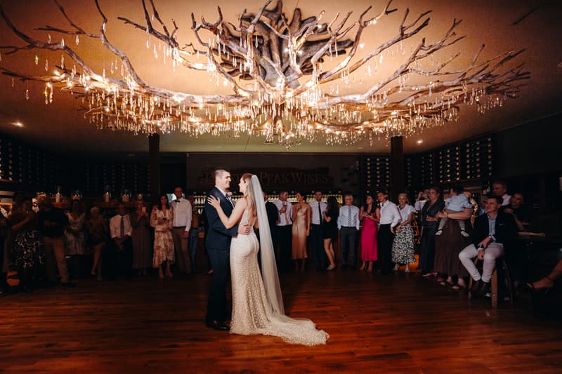 Bride Chelsea and groom Brent share a dance on the wooden floor at Preston Peak Winery Functions Centre, surrounded by guests watching under a large decorative chandelier.