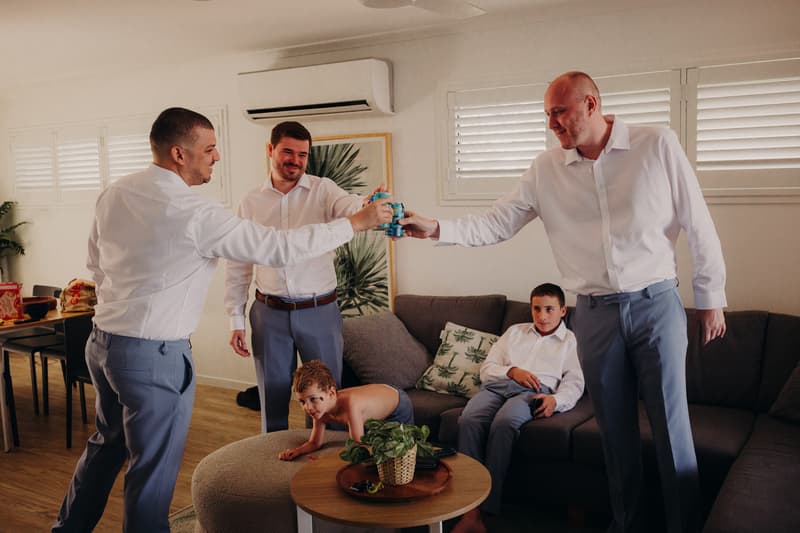 Three men in white shirts and blue pants toast with cans in a living room at Sandstone Point Hotel, while two boys, one shirtless and crawling on an ottoman and the other seated on a couch, watch.