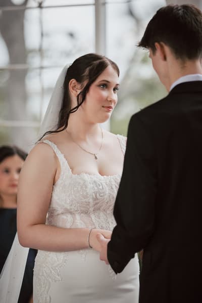 The bride Terion and groom Tyler hold hands facing each other during their wedding ceremony, with a bridesmaid visible in the background. Brisbane wedding videography by She Said Yes.
