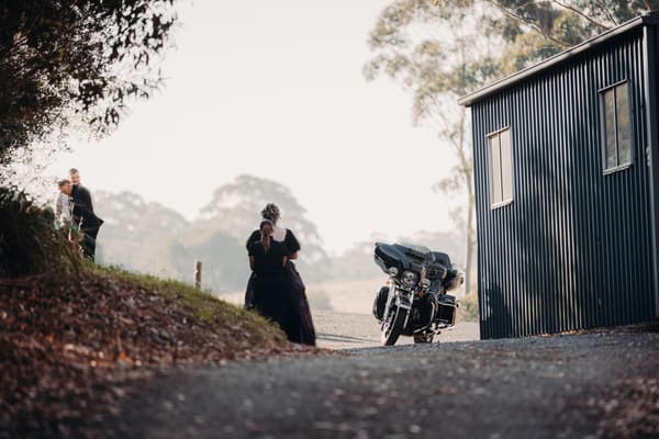Two women in dark dresses stand near a black motorcycle parked beside a dark corrugated metal building at Ocean View Estates — On The Lake, while two men in suits stand on a grassy slope nearby.