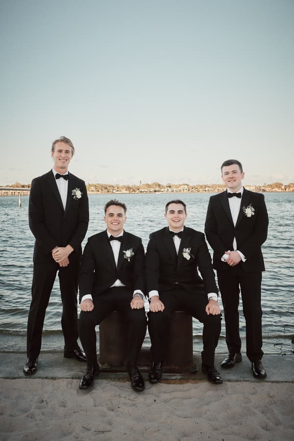 Four groomsmen in black tuxedos with white boutonnieres pose by the water at Sandstone Point Hotel, two seated on bollards and two standing on sand.