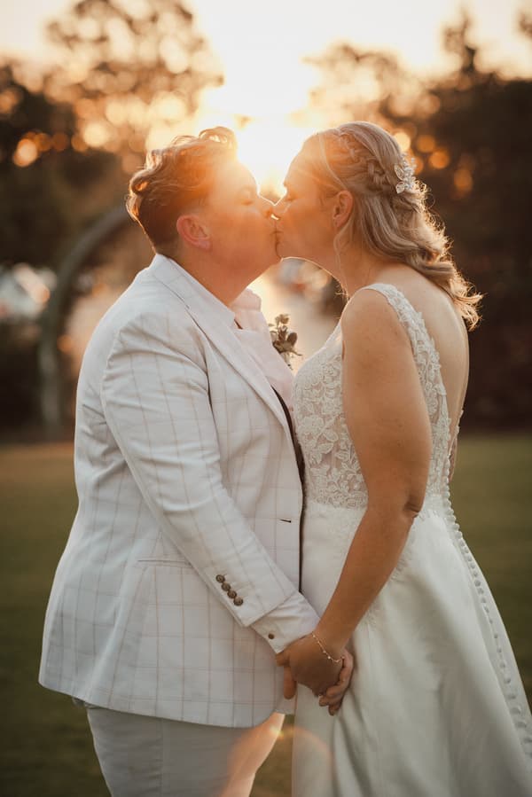 Brides Mandy and Selina share a kiss while holding hands outdoors at Sandstone Point Hotel during their couple portraits session at sunset.