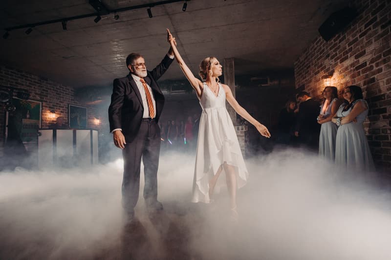 The bride dances with an older man, likely her father, on the fog-covered dance floor at Sandstone Point Hotel — Cellar, while bridesmaids and guests watch from the side.