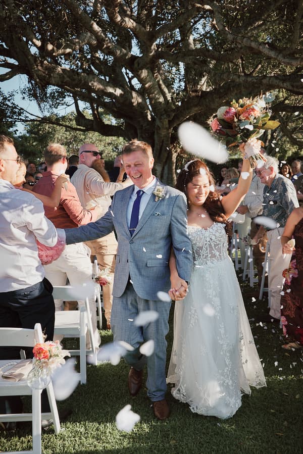 Bride Jheng and groom Dane walk down the aisle holding hands at Sandstone Point Hotel — The Pavilion, surrounded by guests throwing flower petals.
