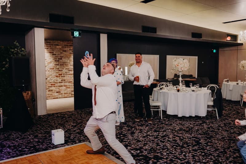 A male guest at the Sandstone Point Hotel — Pumicestone Room reception stage reaches up to catch a thrown object while other guests watch nearby.