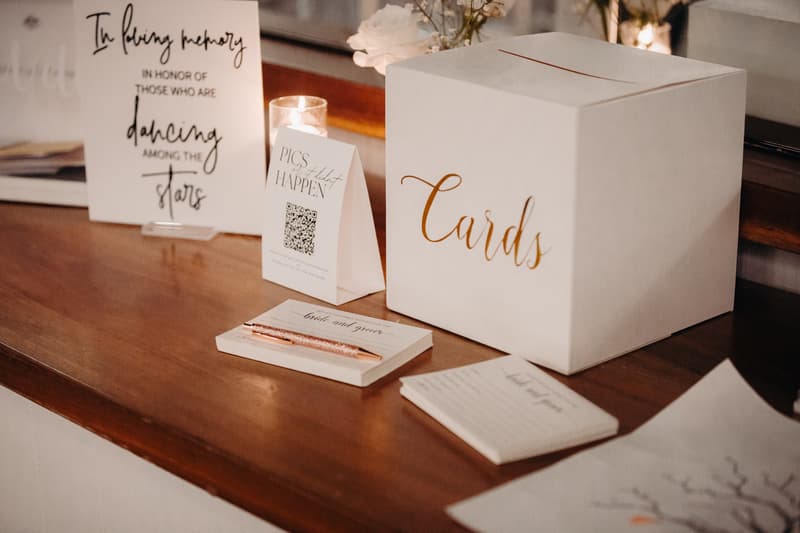A wedding card box with the word 'Cards' in gold script sits on a wooden surface at Sandstone Point Hotel — Pumicestone Room, accompanied by a candle, a sign reading 'In loving memory in honor of those who are dancing among the stars', a 'Pics or it didn't happen' sign with a QR code, and guest book pads labeled 'bride and groom'.