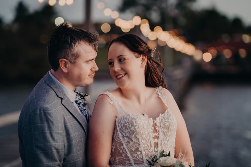 Rebecca the bride and Dale the groom pose together on a wooden pier at Sandstone Point Hotel with string lights blurred in the background over water.