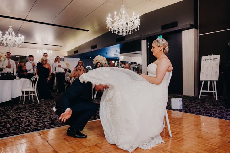 The groom Dylan crouches in front of the seated bride Emily, lifting her wedding dress during the reception at Sandstone Point Hotel — Pumicestone Room, while guests watch and smile in the background.