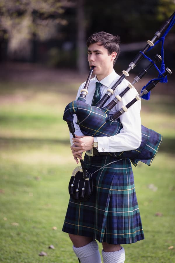 A bagpiper in traditional Scottish attire plays the bagpipes outdoors on grass at Toowong Rowing Club.