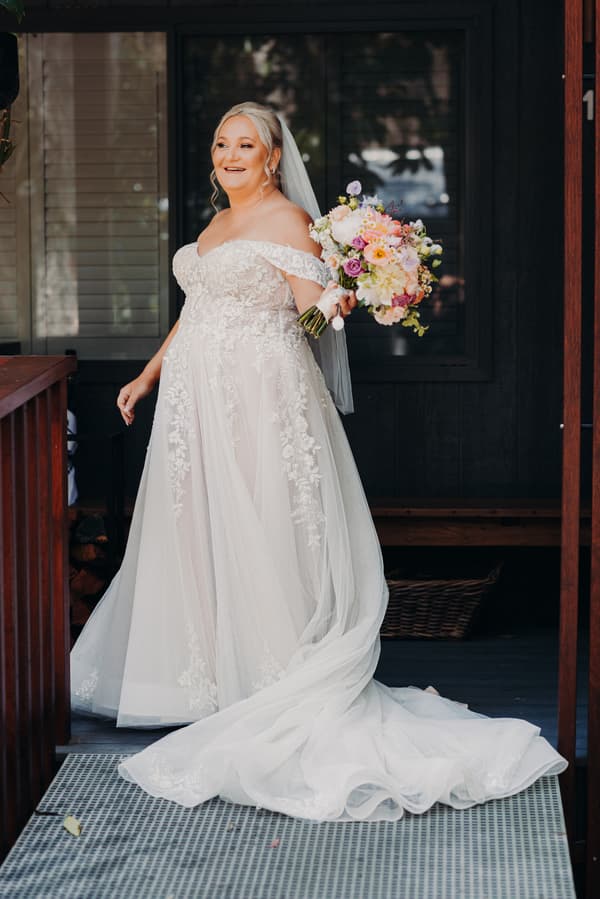 The bride Chantelle stands holding a bouquet of flowers wearing an off-the-shoulder lace wedding gown with a long train and veil at Sandstone Point Hotel.