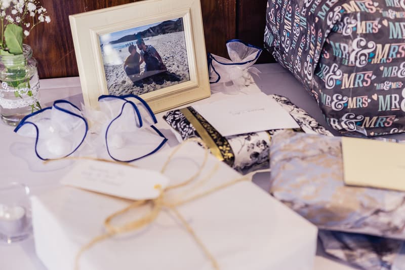 A table at the reception in The Malouf Room at Toowong Rowing Club displaying wrapped wedding gifts, a framed photo of a couple on a beach, small decorative white fabric flowers, and a glass jar with baby's breath flowers.