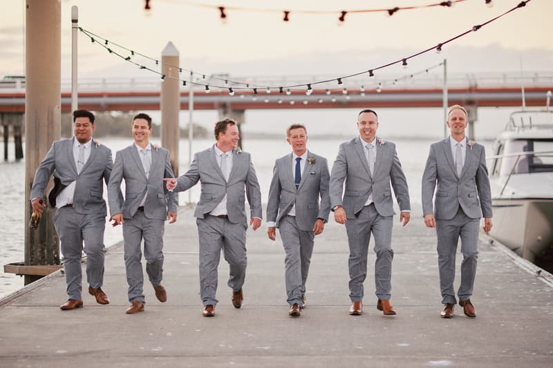 Six groomsmen in grey suits walk along a pier at Sandstone Point Hotel, with water and a boat visible in the background.