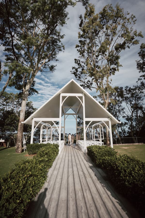 Wedding ceremony setup at Sandstone Point Hotel — The Pavilion with a white open-air structure, wooden aisle, and a small group of people gathered near the altar.
