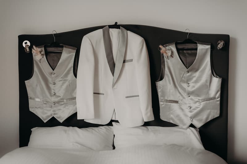 Two silver vests with boutonnieres and a white tuxedo jacket with grey lapels hanging on the headboard of a bed at Royal on the Park.