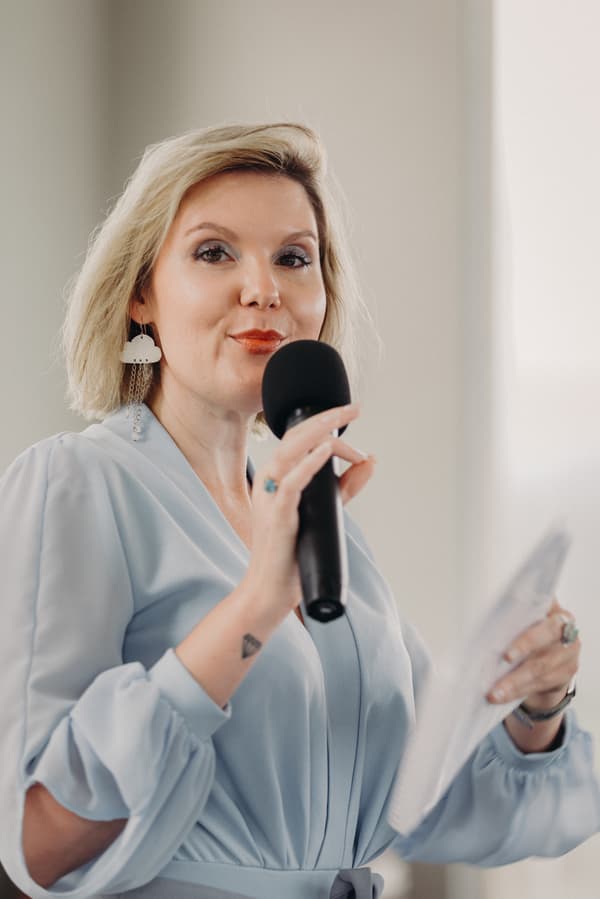 A woman in a light blue dress holds a microphone and a piece of paper, speaking indoors at Preston Peak Winery Functions Centre.
