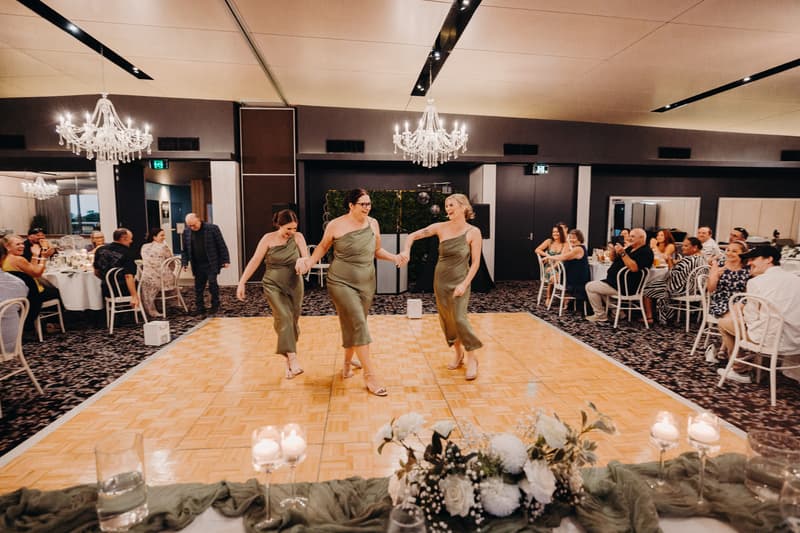 Three bridesmaids in olive green dresses hold hands and dance on the dance floor at the Sandstone Point Hotel — Pumicestone Room during the wedding reception, while guests seated at round tables watch and smile.