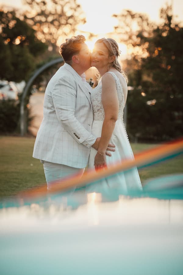 Mandy and Selina share a tender moment during their couple portraits at Sandstone Point Hotel, with Selina kissing Mandy on the cheek as the sun sets behind them.