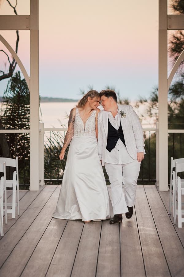 Mandy in a white wedding gown and Selina in a white suit with a black vest walk closely together at Sandstone Point Hotel — The Pavilion with a waterfront view in the background.