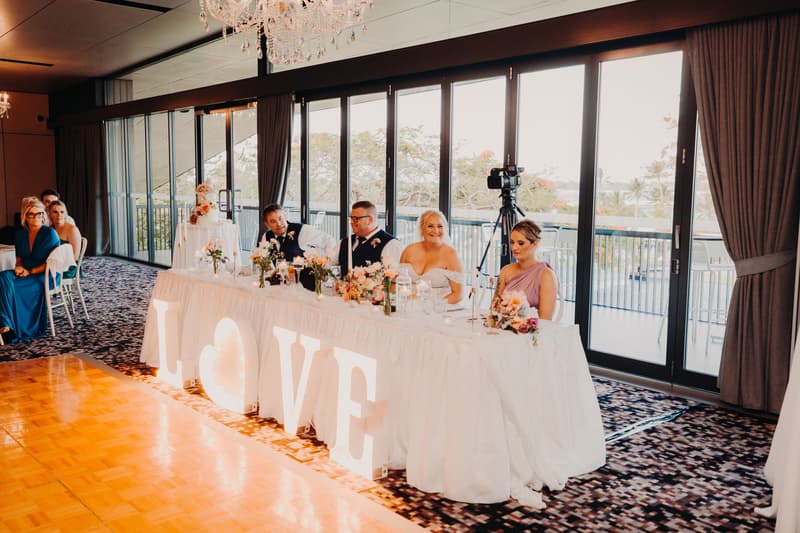 The bride Chantelle and groom Peter sit at the head table in the Pumicestone Room at Sandstone Point Hotel during the wedding reception, accompanied by two guests. The table is decorated with floral arrangements and large illuminated letters spelling LOVE.