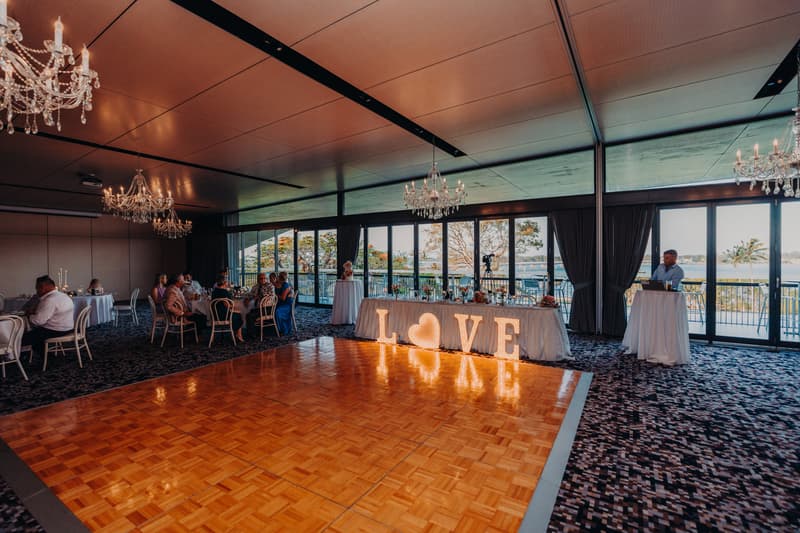 Reception room at Sandstone Point Hotel — Pumicestone Room with guests seated at tables, a lit 'LOVE' sign on a table near the windows, and a man standing at a high table near the glass doors overlooking the water.