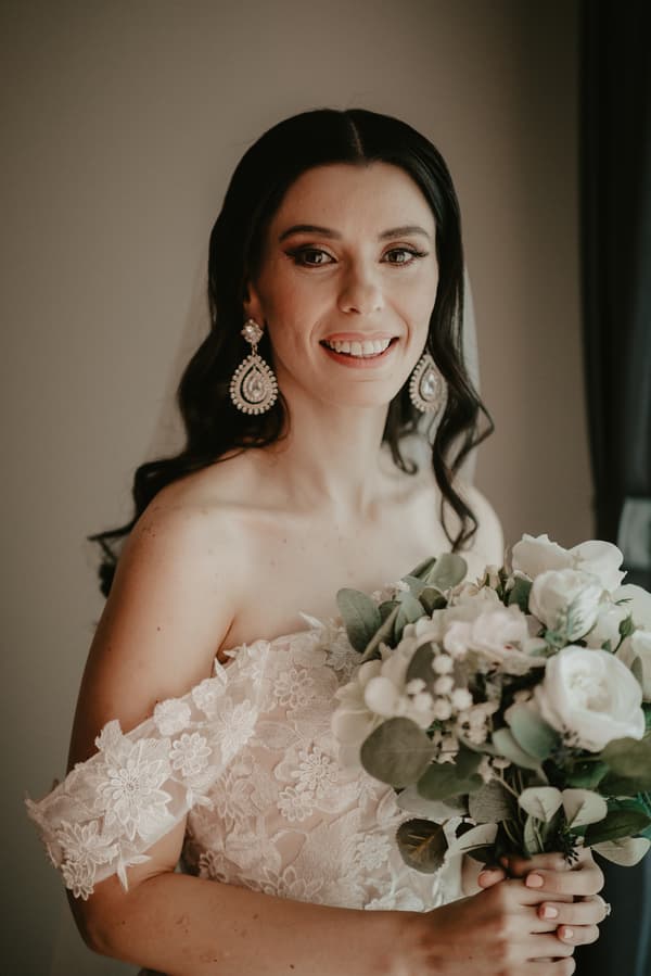 A bride with long dark hair wearing large decorative earrings and an off-shoulder lace wedding dress holds a bouquet of white flowers and greenery.