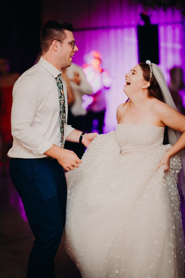 Bride Lilly and groom Connor dance together at the reception in The Shed at Yabbaloumba Retreat, with Lilly holding up her wedding dress and both smiling.
