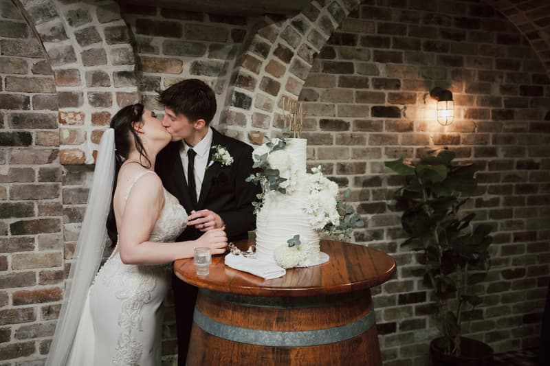 Terion and Tyler kiss beside their wedding cake on a wooden barrel table at Sandstone Point Hotel — The Cellar, with a brick wall and wall-mounted light in the background.