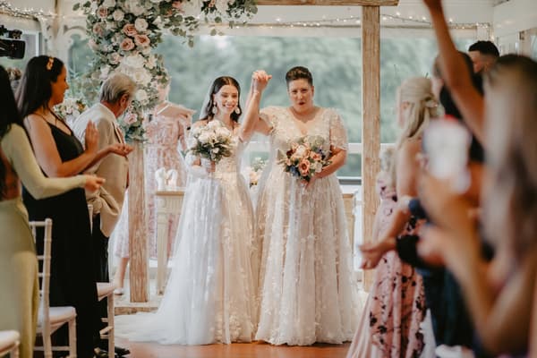 Two brides in white lace wedding dresses holding bouquets stand together holding hands raised, surrounded by applauding guests in a decorated indoor venue with floral arch and wooden beams.