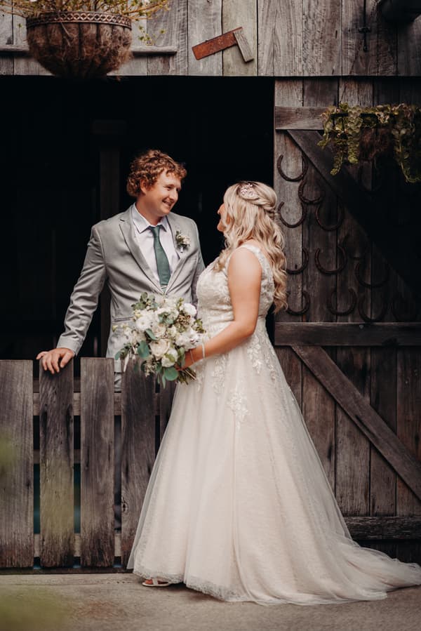 Bride Chloe in a white lace wedding gown holding a bouquet and groom Brodie in a light grey suit with a green tie stand facing each other by a rustic wooden fence and barn door at Sandstone Point Hotel.