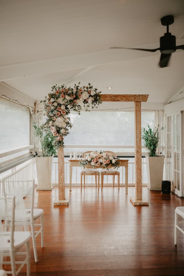 Empty wedding ceremony setup with a wooden arch decorated with flowers, a small table with floral arrangements, two chairs, and white chairs arranged for guests in a bright indoor space.