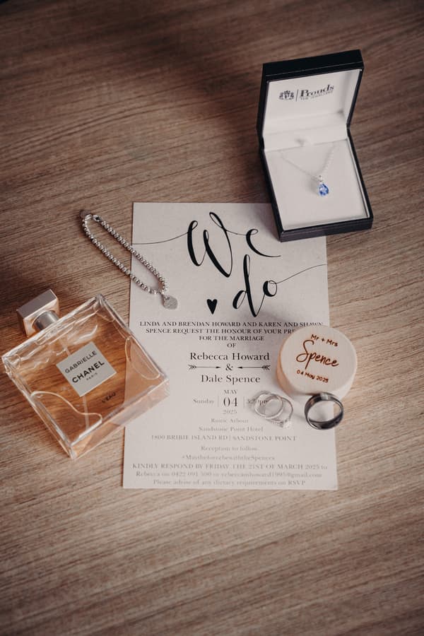 Wedding invitation for Rebecca Howard and Dale Spence's marriage at Sandstone Point Hotel Rustic Arbour on May 4, 2025, displayed with a Chanel Gabrielle perfume bottle, a silver bracelet, a necklace with a blue pendant in a box, and two wedding rings on a wooden surface.